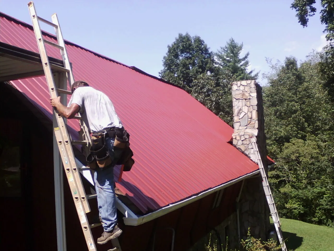 Roofer on a ladder beside a red metal roof and stone chimney — Soffit & Fascia Repair by Roofing Brothers in Fontanelle